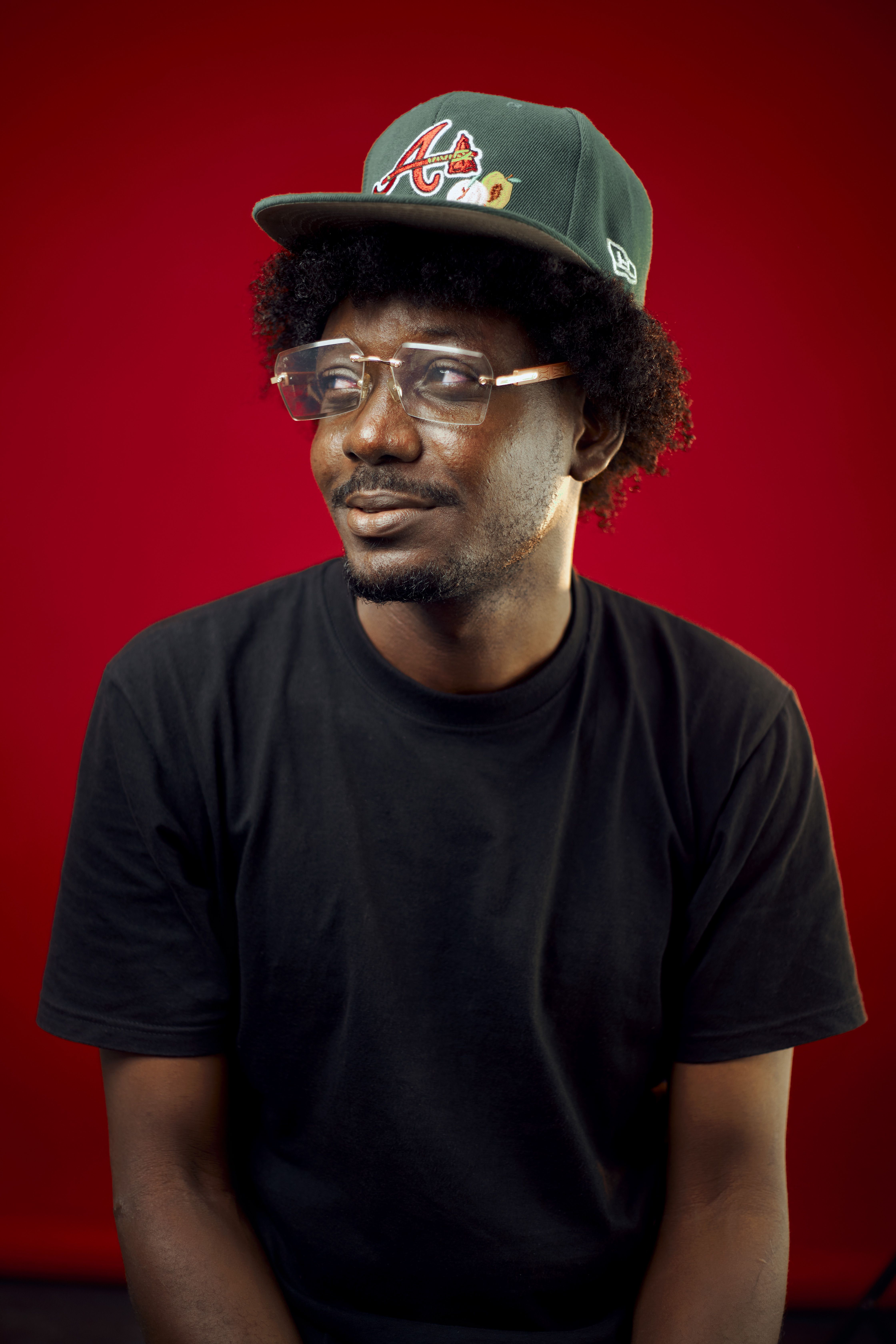 Casual portrait of a man wearing a hat and glasses against a red backdrop.