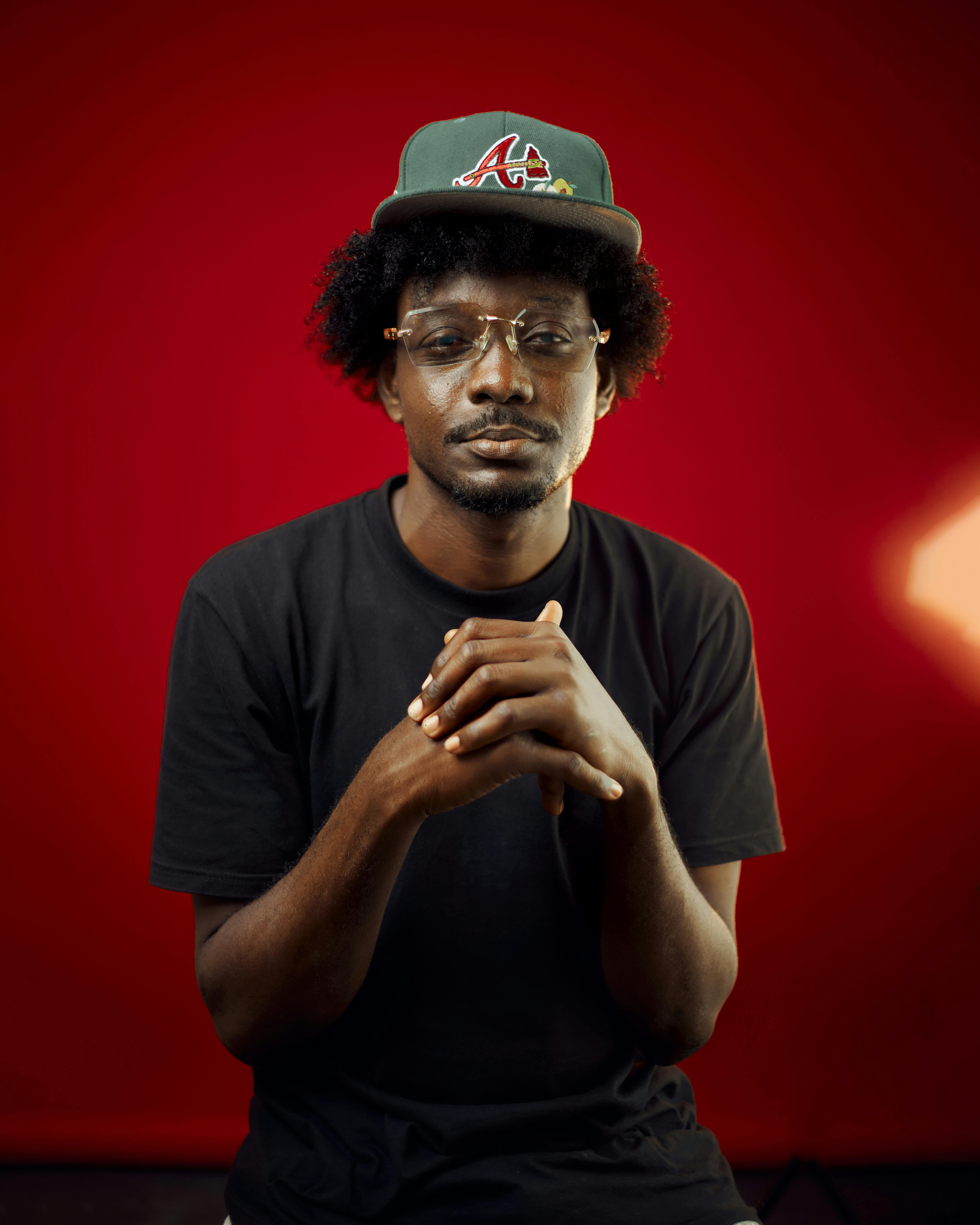 Thoughtful portrait of a man in glasses and baseball cap against a vibrant red backdrop.