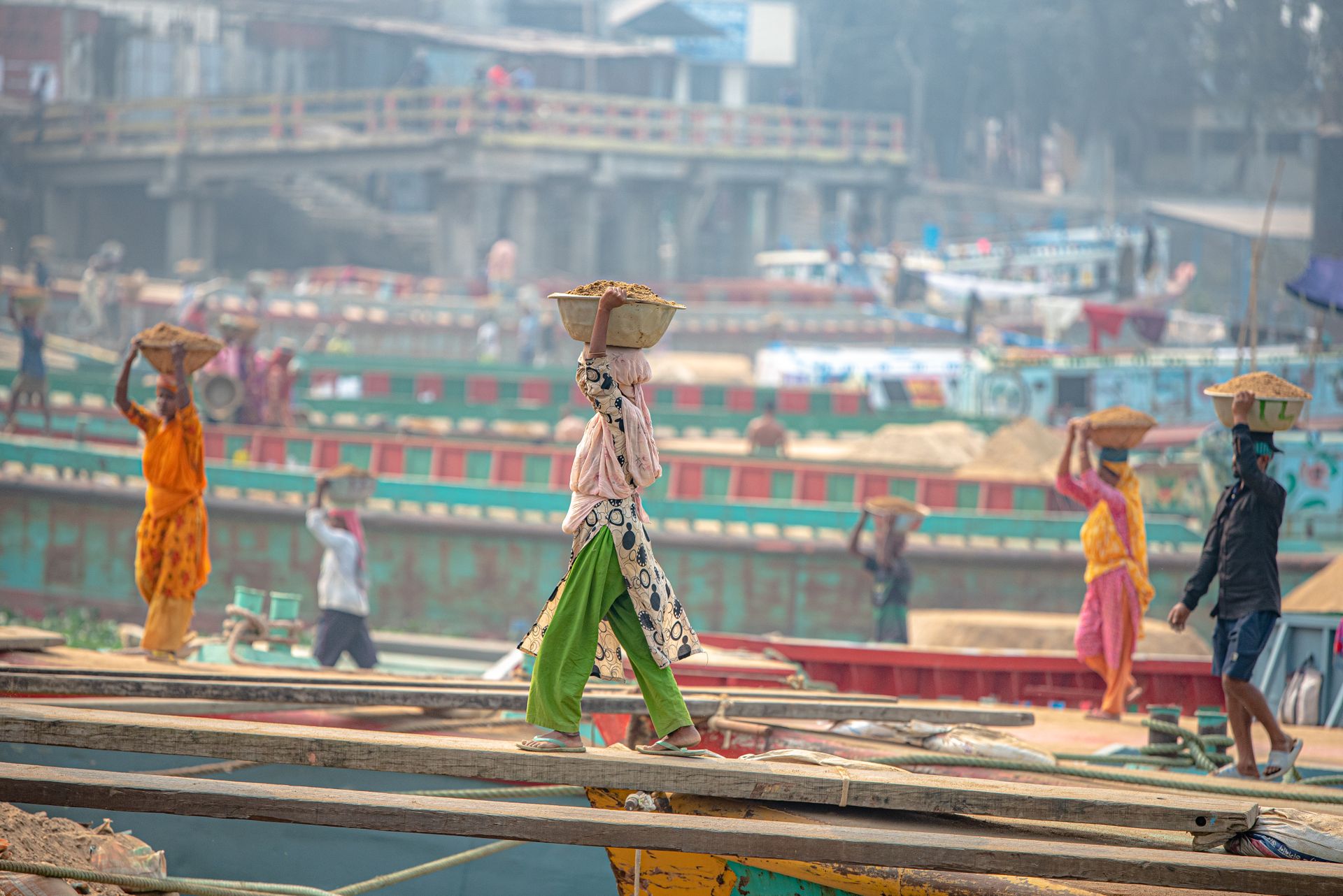 Laborers transport sand at a busy river port, showcasing vibrant cultural attire and activity.