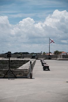 A captivating view of an ancient fort exterior under a cloudy sky, featuring benches and a flag.