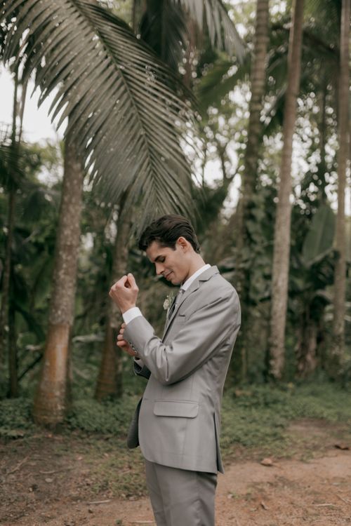 Free A young man in a gray suit adjusting his cufflinks in an outdoor setting surrounded by tropical trees. Stock Photo