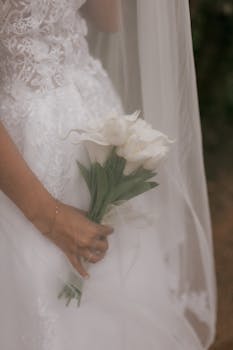 Close-up of a bride holding a bouquet of white tulips with a lace wedding dress background.