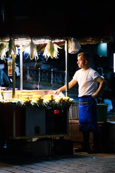A street vendor selling roasted corn under bright lights in a night market setting.