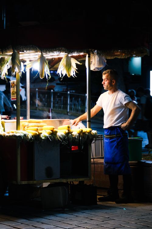 Free A street vendor selling roasted corn under bright lights in a night market setting. Stock Photo