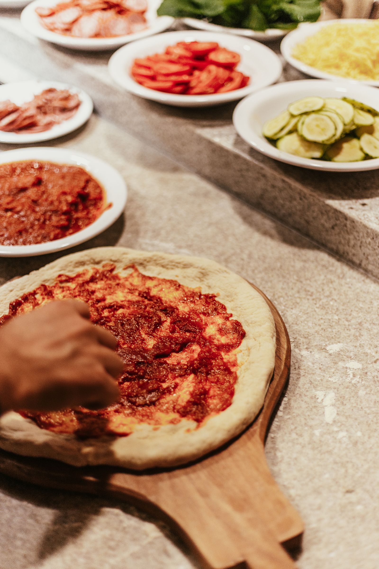 Mani che stendono il pomodoro su una base, accanto a zucchine, salumi e formaggio grattugiato su un banco di pietra.