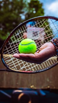 A close-up view of a tennis ball held in hand with a racket background, set outdoors.