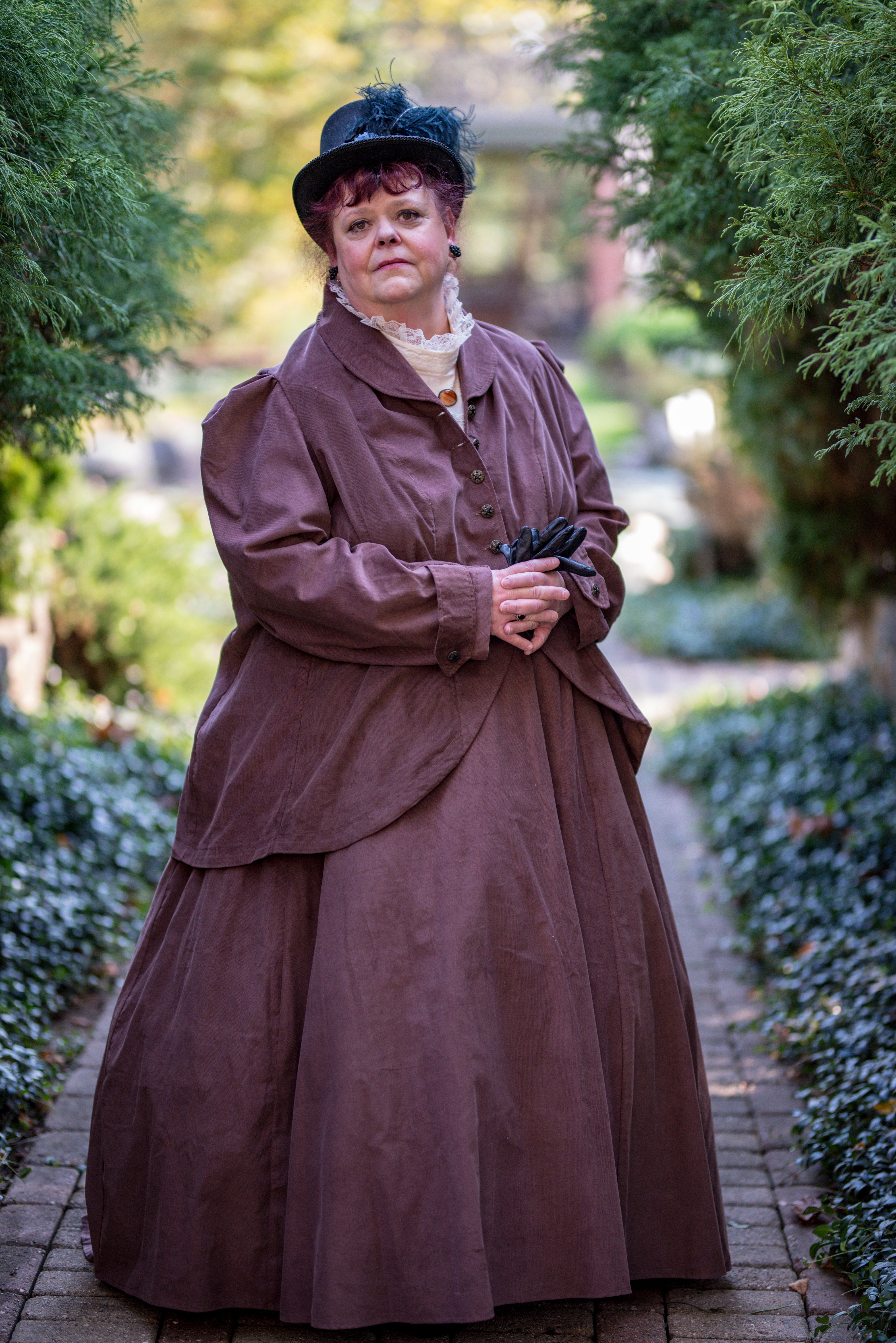 Woman in Victorian dress poses in lush garden pathway, embodying historical elegance.