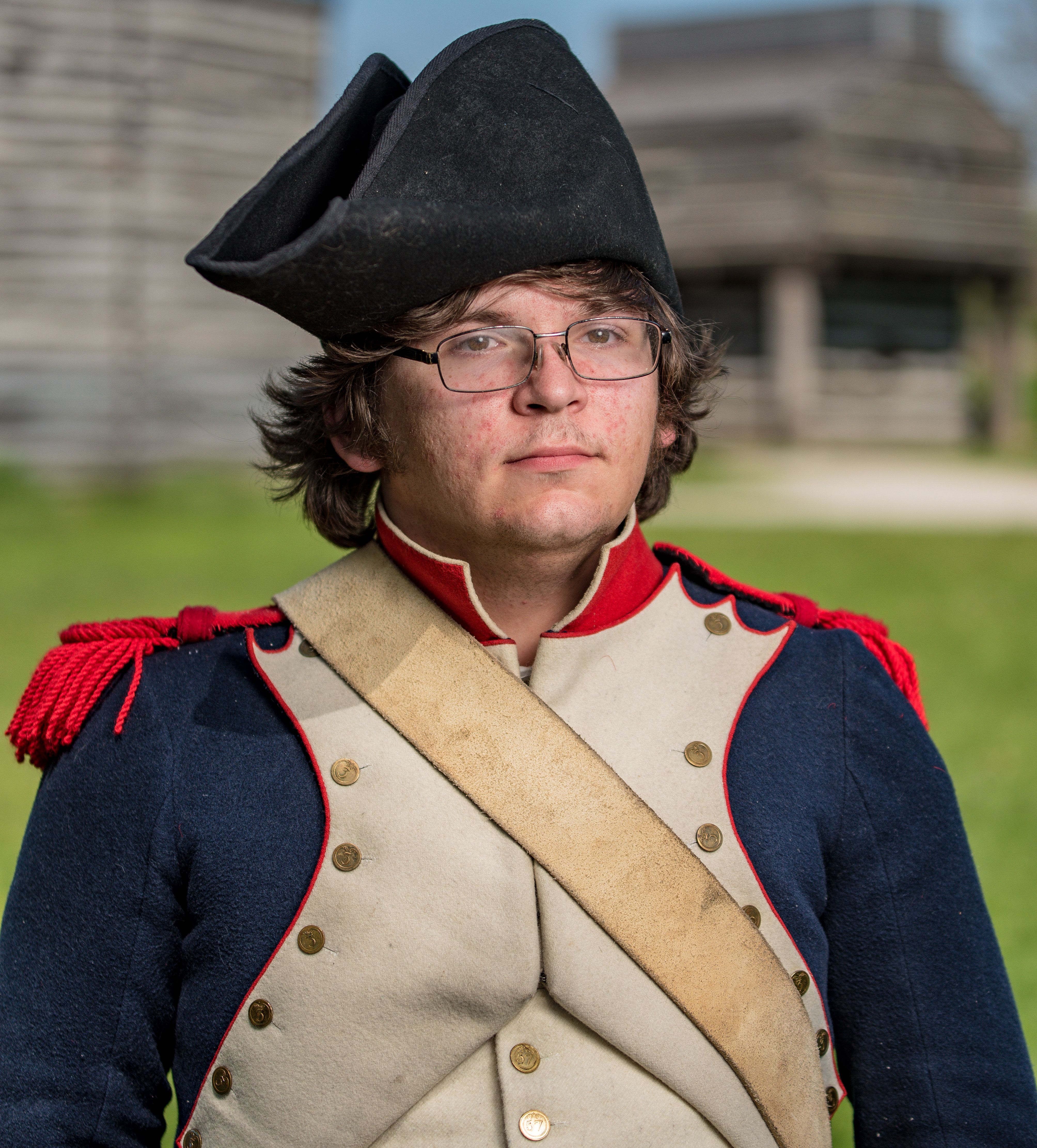 A young man in historical military attire poses outdoors with a rustic backdrop.