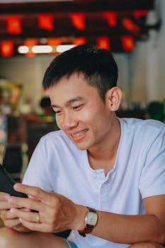 A young man smiles while looking at his smartphone inside a cozy room.