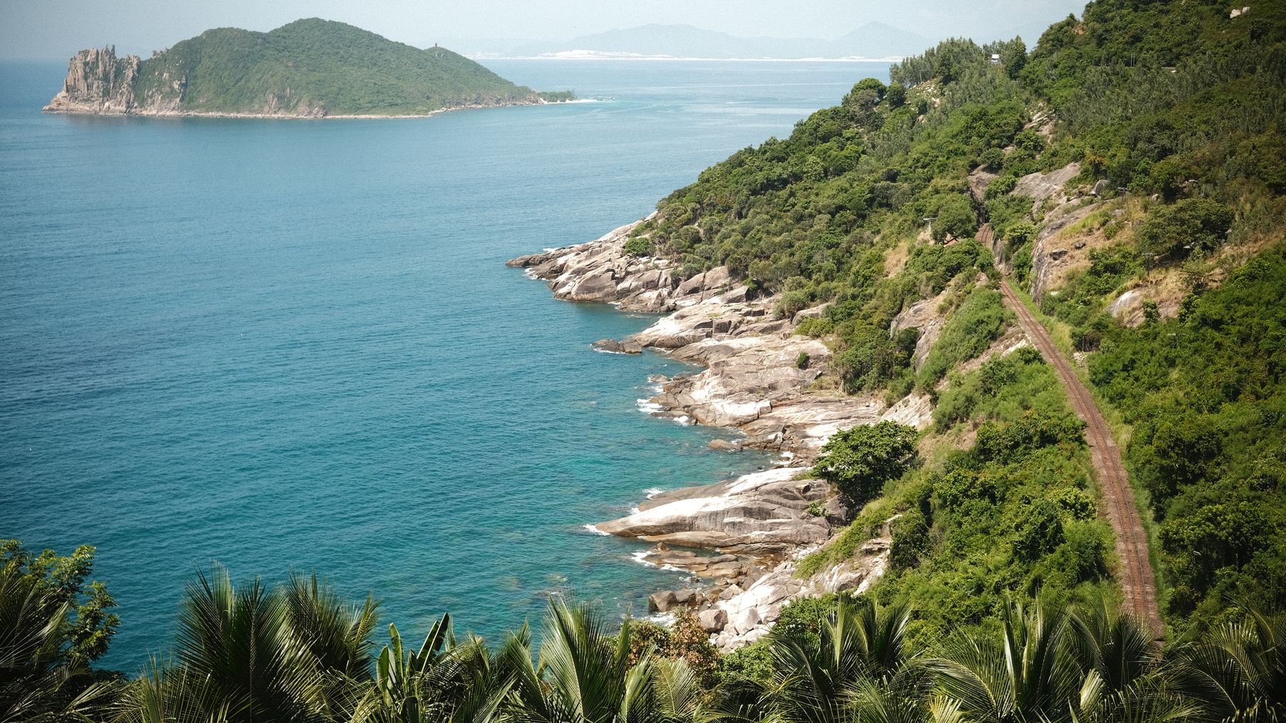 A breathtaking view of a coastal path with lush greenery and a distant island in bright daylight.