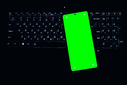 A smartphone with a bright green screen rests on a backlit keyboard, creating a striking contrast in a dark setting.