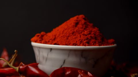 A bowl of vibrant red chili powder surrounded by whole dried chilies on a dark background.