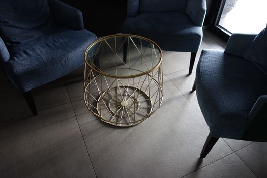 Stylish indoor lounge featuring blue armchairs around a glass-top metal table on tiled flooring.