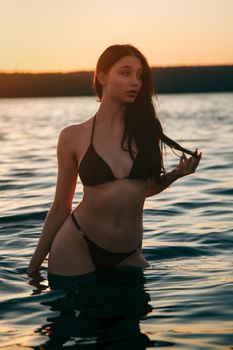 Elegant woman in a bikini standing in calm waters during sunset, embodying serenity and summer vibes.