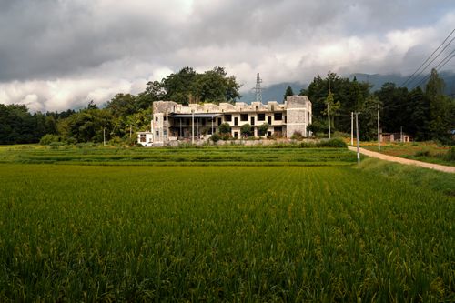 Free An abandoned building surrounded by lush greenery and fields under a cloudy sky. Stock Photo