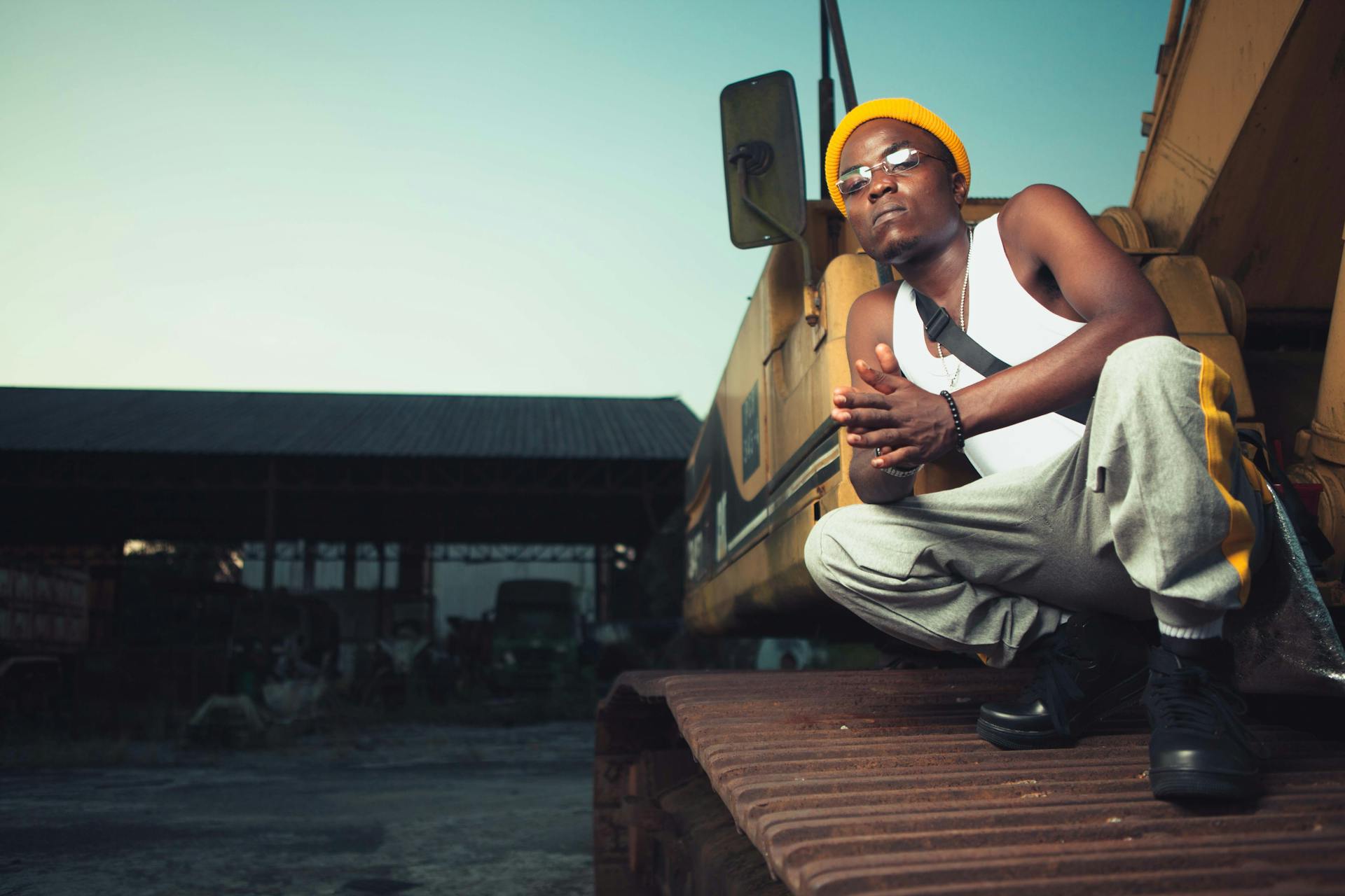 Man in casual attire poses confidently on construction equipment in an industrial setting.