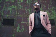 一位时尚男士身着粉色西装外套，戴着太阳镜，在翠绿的乡村背景前自信摆拍，尽显前卫风范