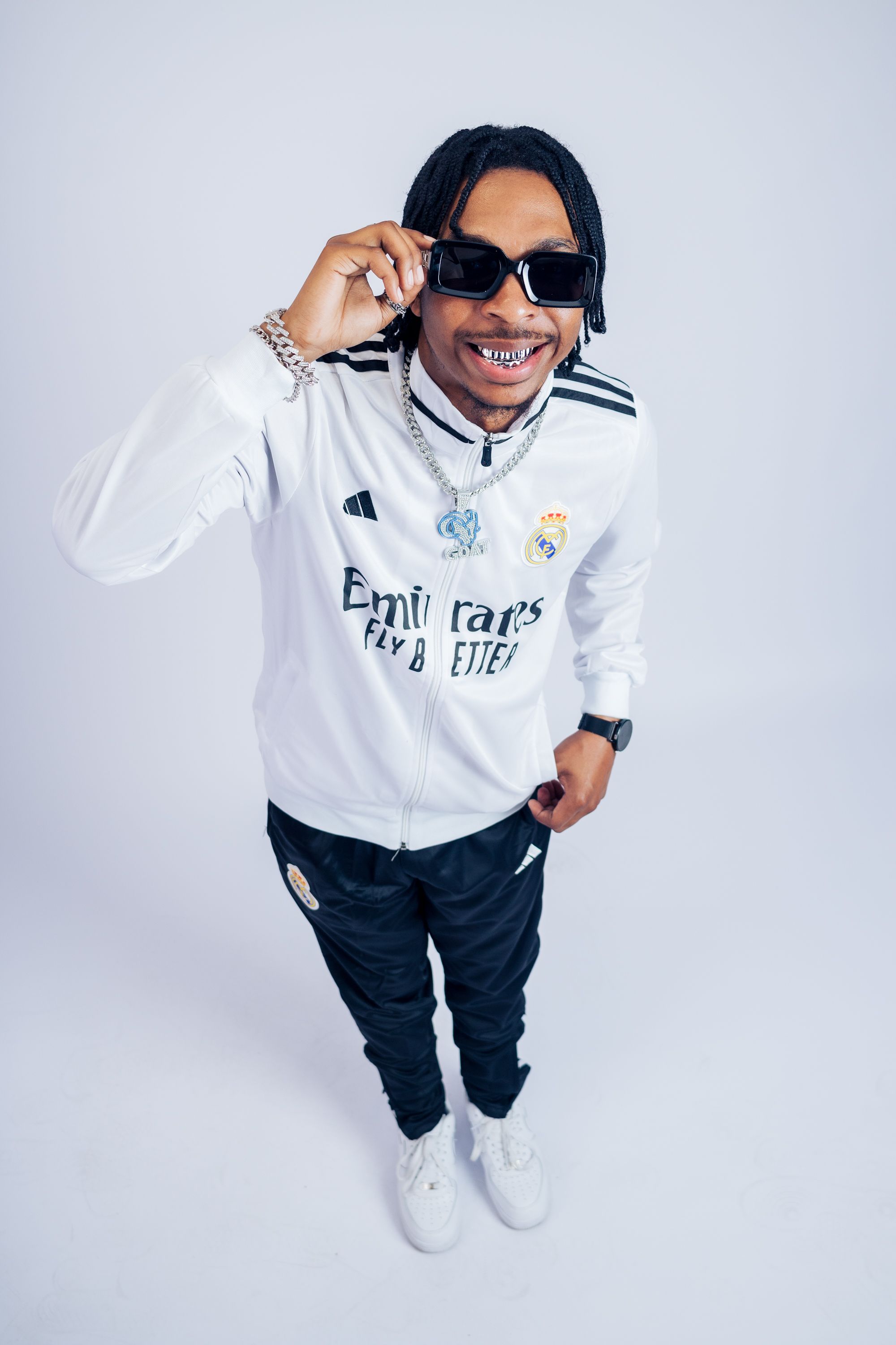 Man in white tracksuit poses confidently wearing sunglasses against white background.