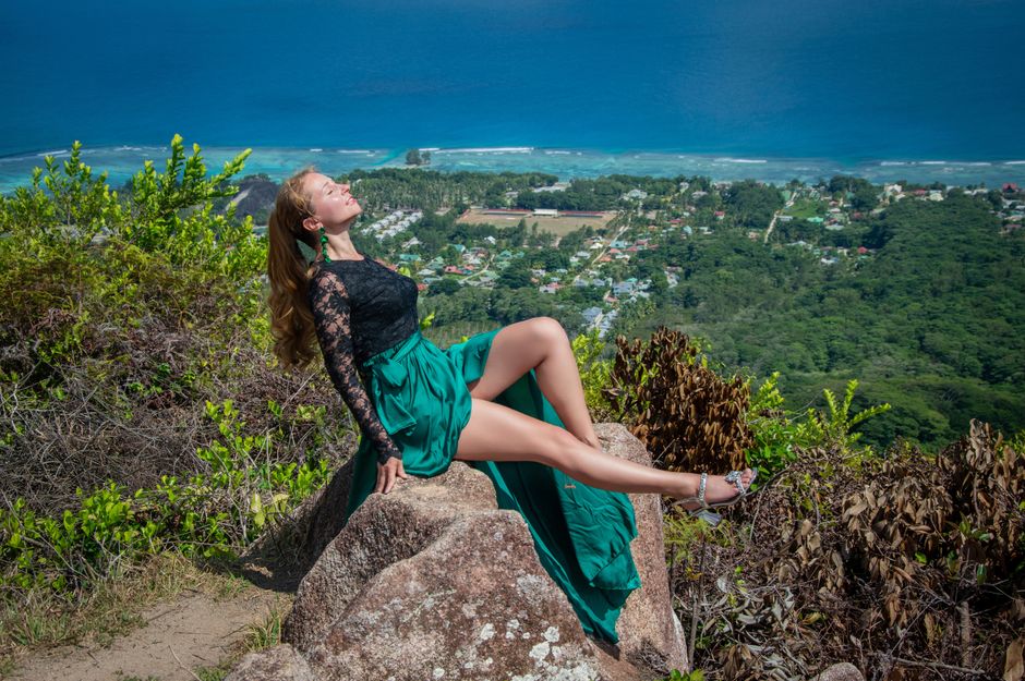 A woman in a green dress relaxing on a cliff with a stunning ocean view in the background.