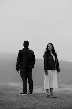 A couple standing apart in a serene outdoor setting, captured in black and white.