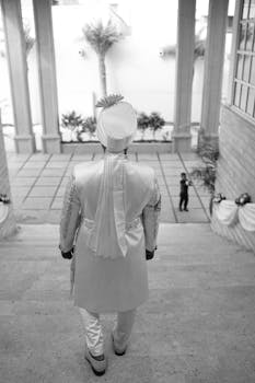 Indian groom in traditional attire walking down a grand staircase, captured in black and white for a classic wedding moment.