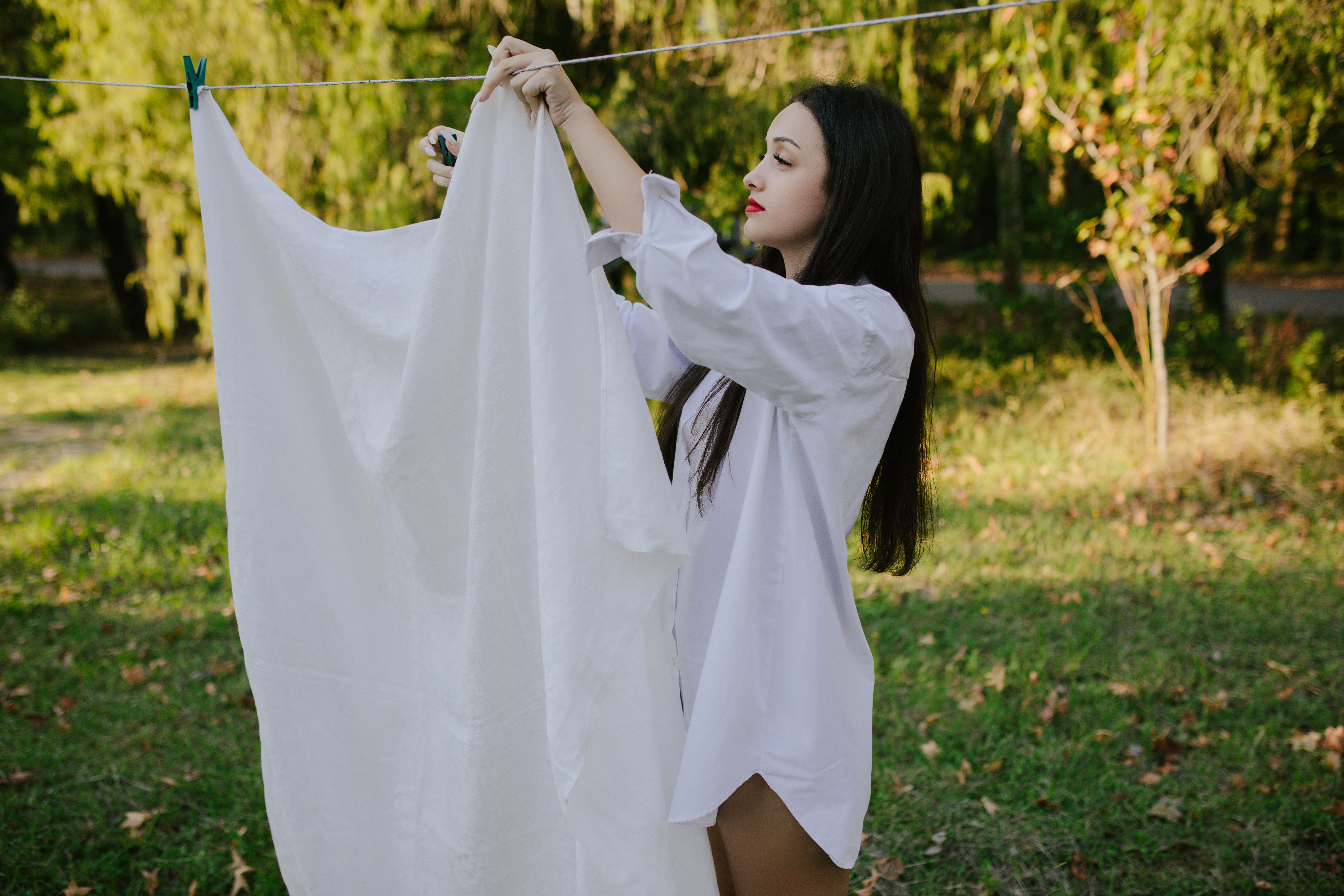 A young woman in a white shirt hangs a sheet outdoors on a sunny day, surrounded by nature.
