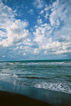 Expansive view of ocean waves crashing on a sandy beach under a broad blue sky with fluffy clouds.