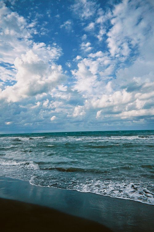 Free Expansive view of ocean waves crashing on a sandy beach under a broad blue sky with fluffy clouds. Stock Photo