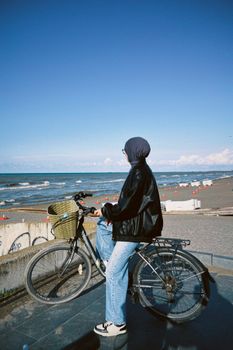 A woman wearing a hijab sits on a bicycle by the ocean, enjoying a sunny beach view.