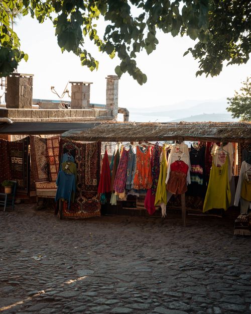 Free Outdoor market stall displaying vibrant textiles and clothes, shaded by leafy trees in a quaint setting. Stock Photo