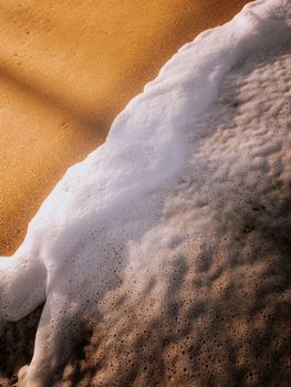 Close-up of sea foam washing over a sandy beach in warm sunlight, offering tranquility and serenity.