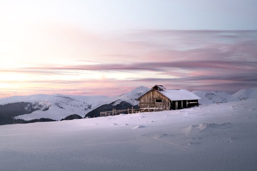 Serene winter landscape with a rustic cabin in the snow during a colorful sunset in Romania.