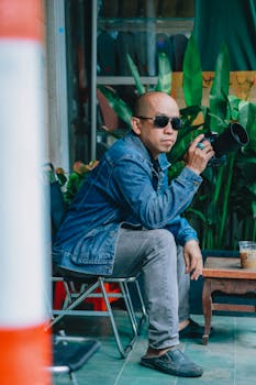 Man with sunglasses holding a camera, sitting outdoors in casual attire.