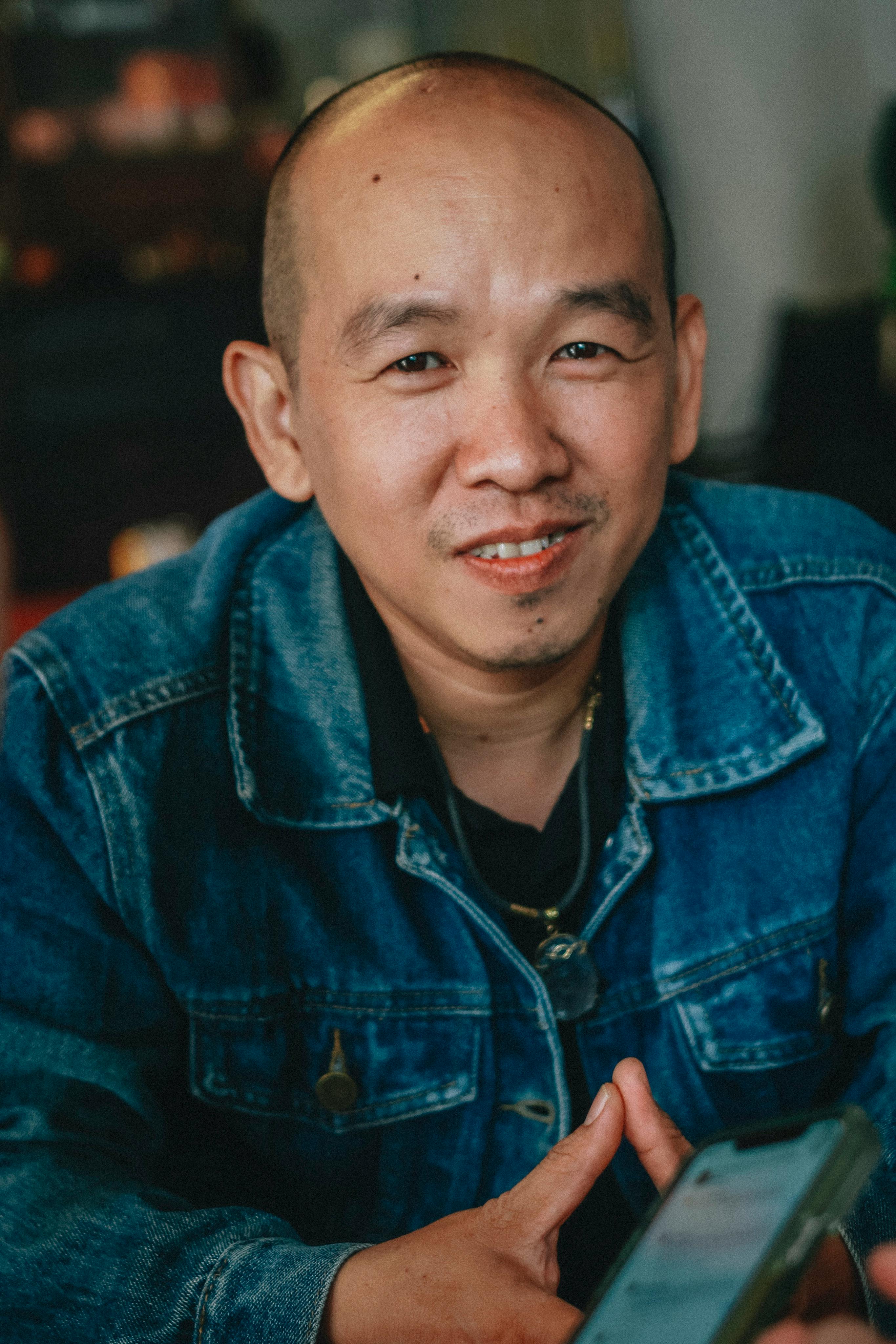 Smiling adult man in denim jacket seated indoors, holding a smartphone, creating a warm and inviting atmosphere.