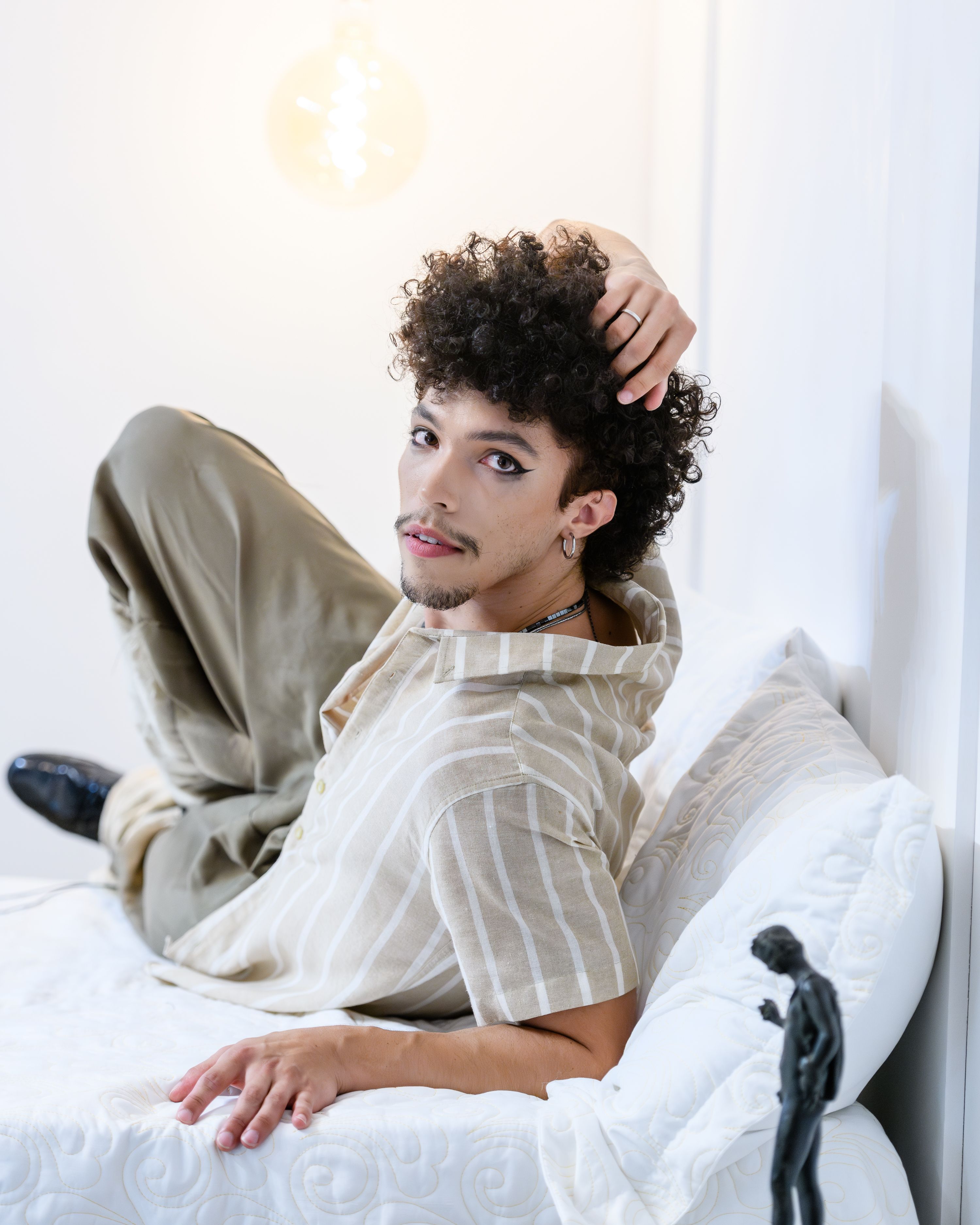 Um jovem de cabelos cacheados relaxa em uma cama dentro de casa, exalando um clima casual e moderno.