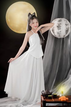 Elegant woman in white gown poses gracefully under a full moon with traditional lantern and tea setup.