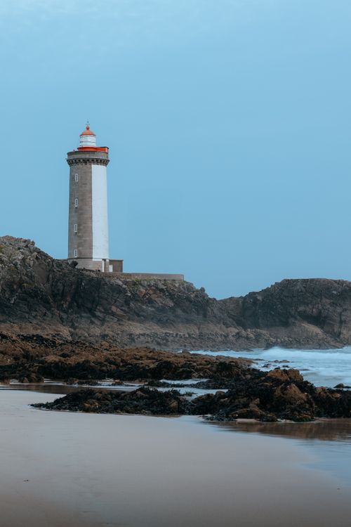 Free A picturesque lighthouse stands majestically on a rocky coast against a blue sky. Stock Photo