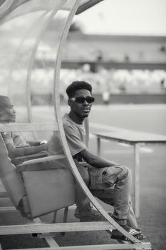 Black and white photo of a young man wearing sunglasses, sitting relaxed on a sports field bench.