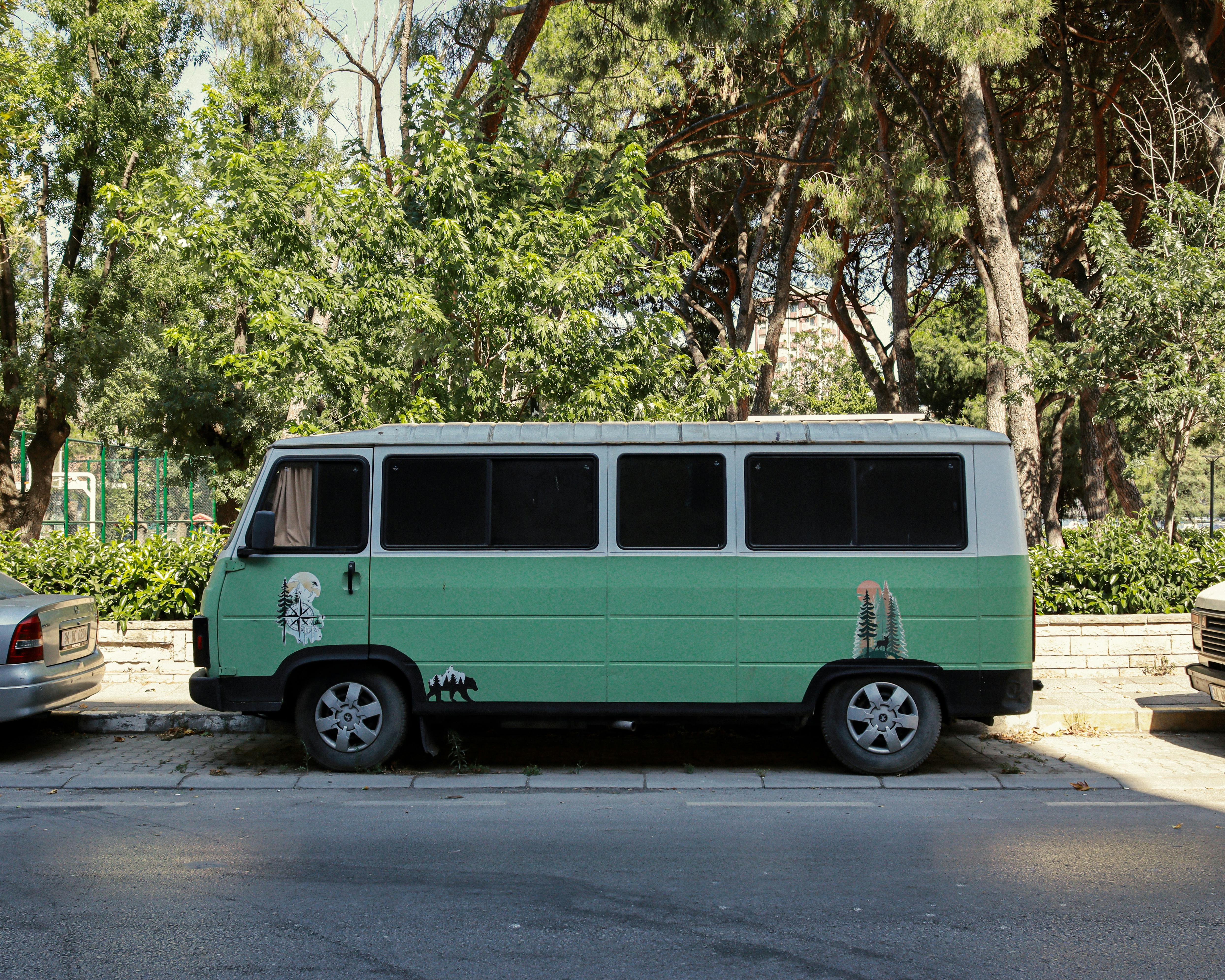 A vintage green camper van parked on a sunny street amidst lush trees in an urban environment.