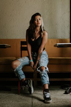 一位穿着破洞牛仔裤和运动鞋的时尚年轻女子，在室内椅子上摆出姿势，展现出休闲别致的风格