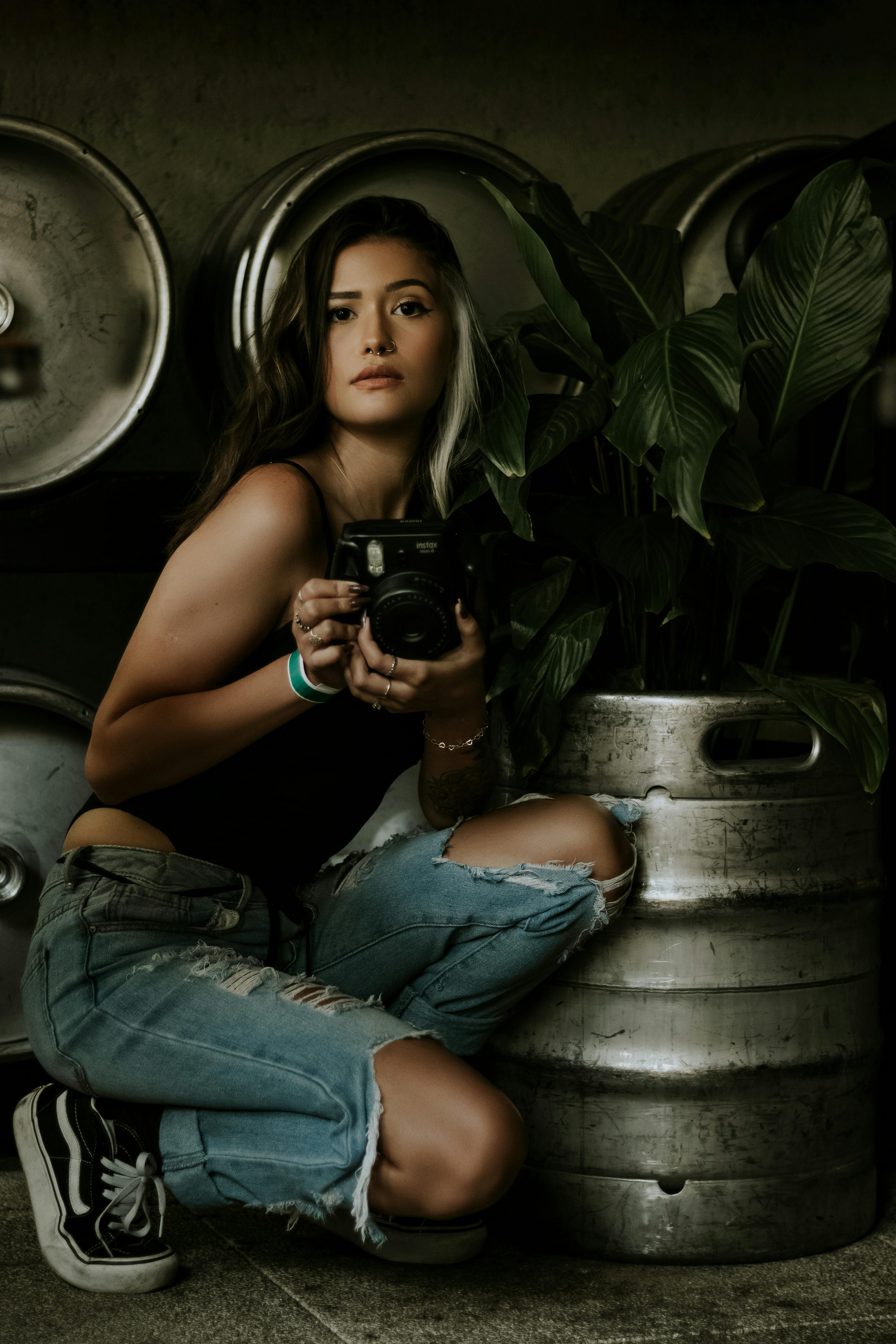 一个女人穿着休闲服，拿着相机，跪在室内的植物和啤酒桶旁