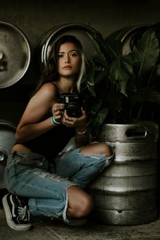 一个女人穿着休闲服，拿着相机，跪在室内的植物和啤酒桶旁