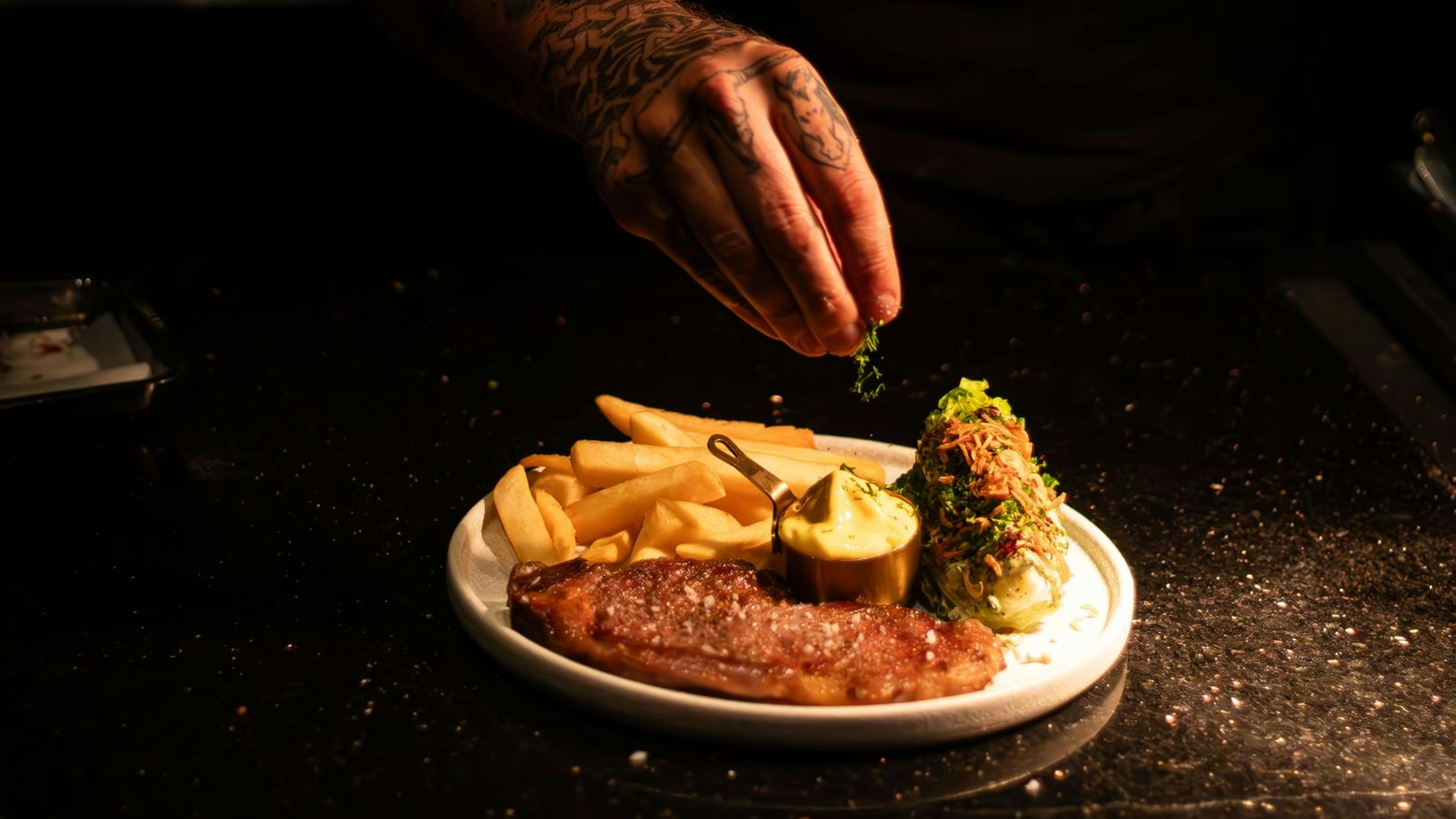 A chef garnishes a steak and fries plate with herbs in a dimly lit restaurant setting.