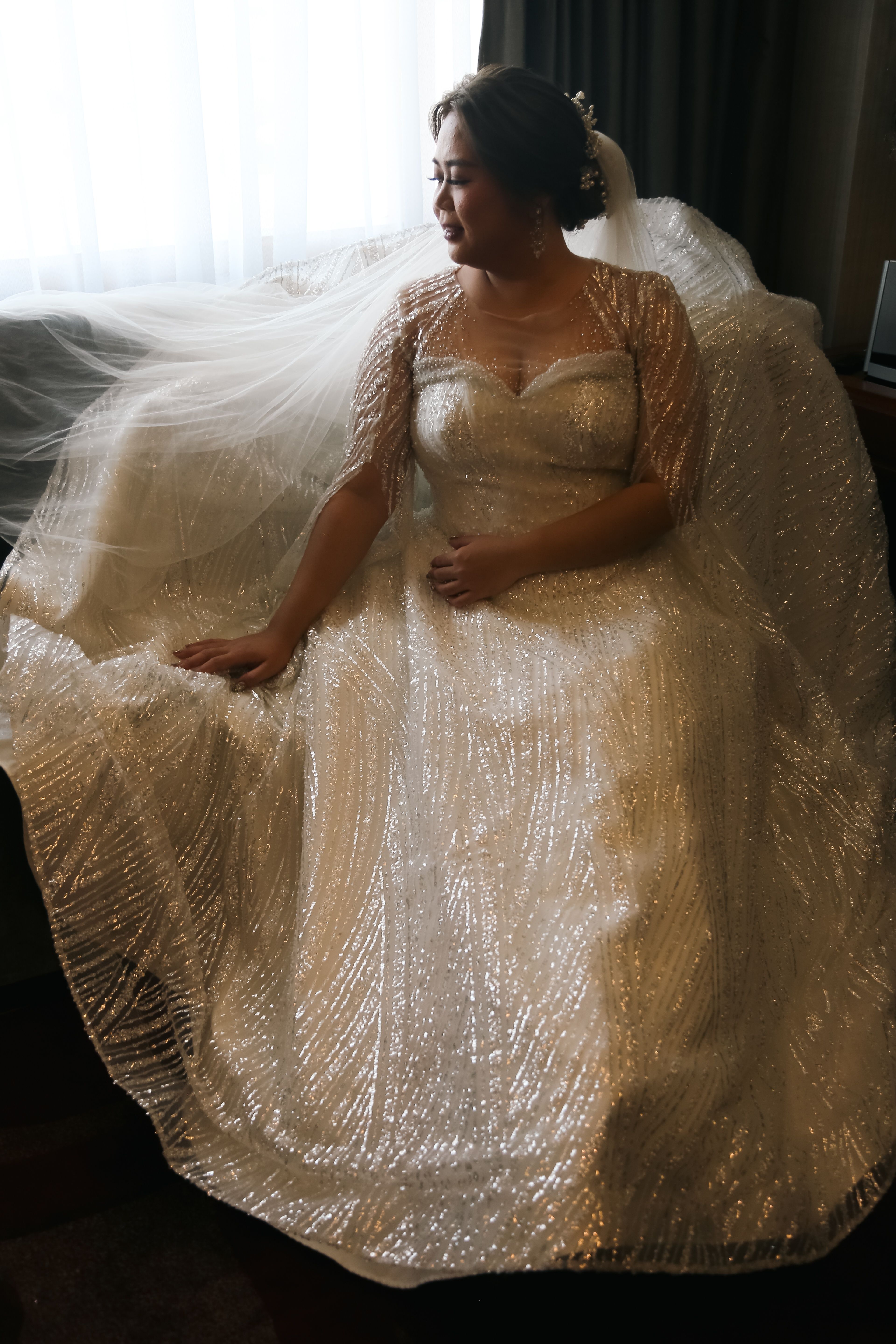 Bride in sparkling wedding dress sitting by window with soft veil.