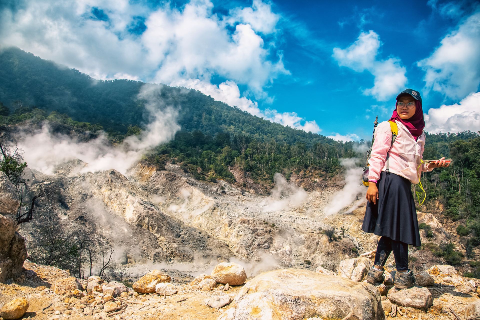 A woman stands in front of a smoking sulfuric volcanic landscape under a bright blue sky.
