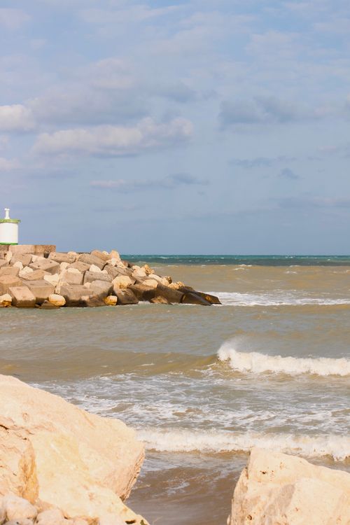 Free Sunlit view of a rocky breakwater and small lighthouse by a wavy seaside, ideal for beach and travel themes. Stock Photo