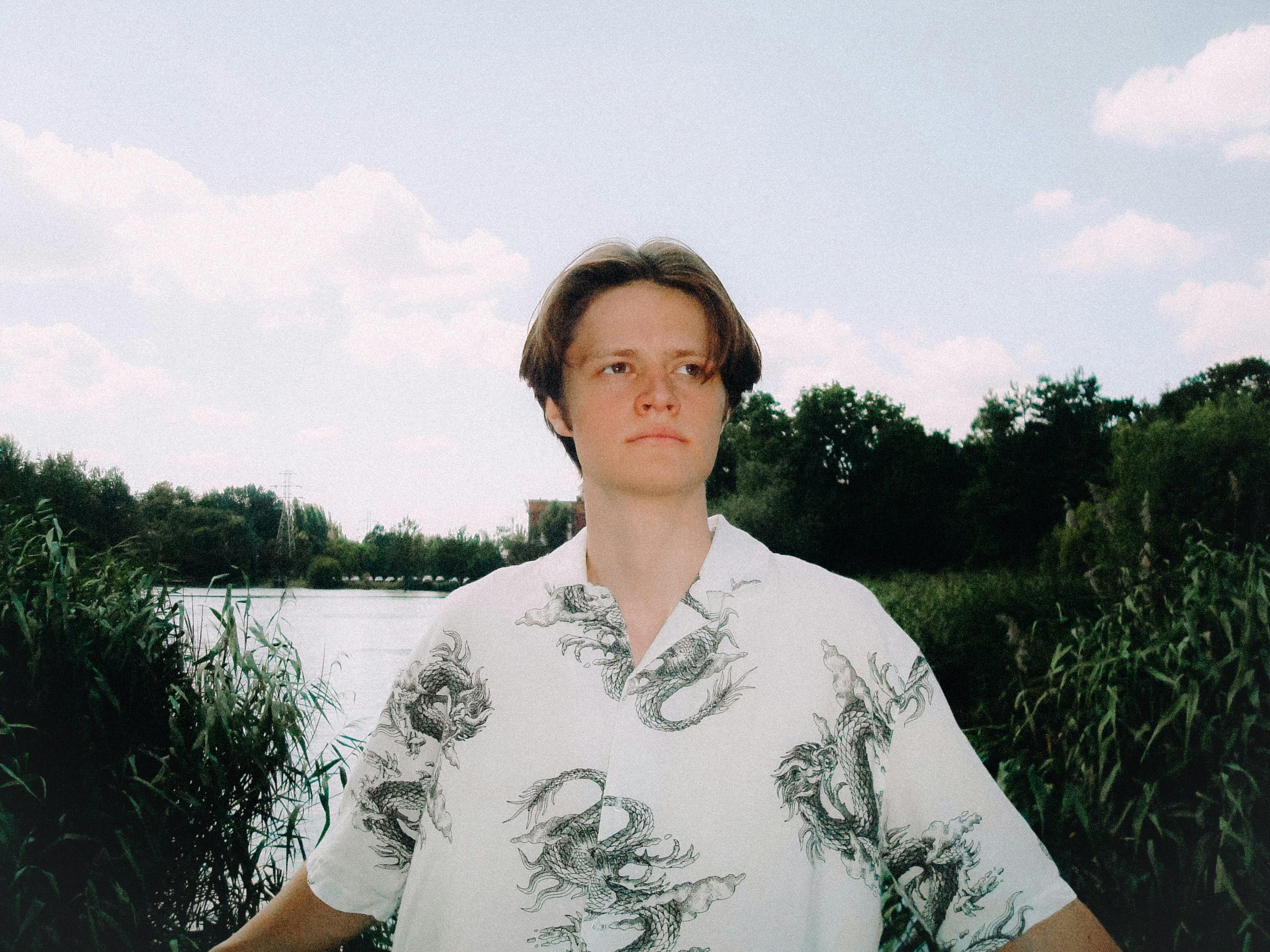 бесплатная Портрет молодого человека в рубашке с драконом, стоящего на берегу пышного озера. Стоковое фото