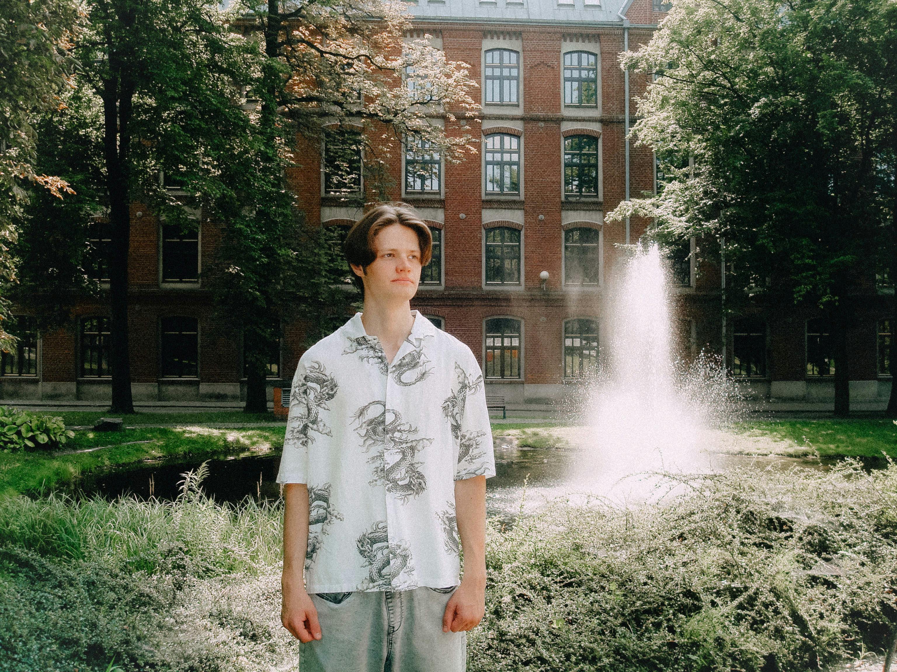 Ilmainen Nuori mies seisoo puutarhassa, taustalla suihkulähde ja historiallinen rakennus. Kuvapankkikuva