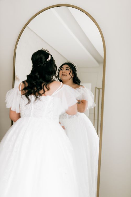 Free A bride in an elegant white dress smiles as she looks at her reflection in a full-length mirror. Stock Photo