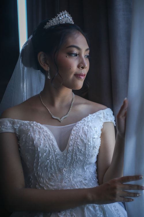 Free Elegant bride in a white gown looking out of the window, creating a serene moment. Stock Photo